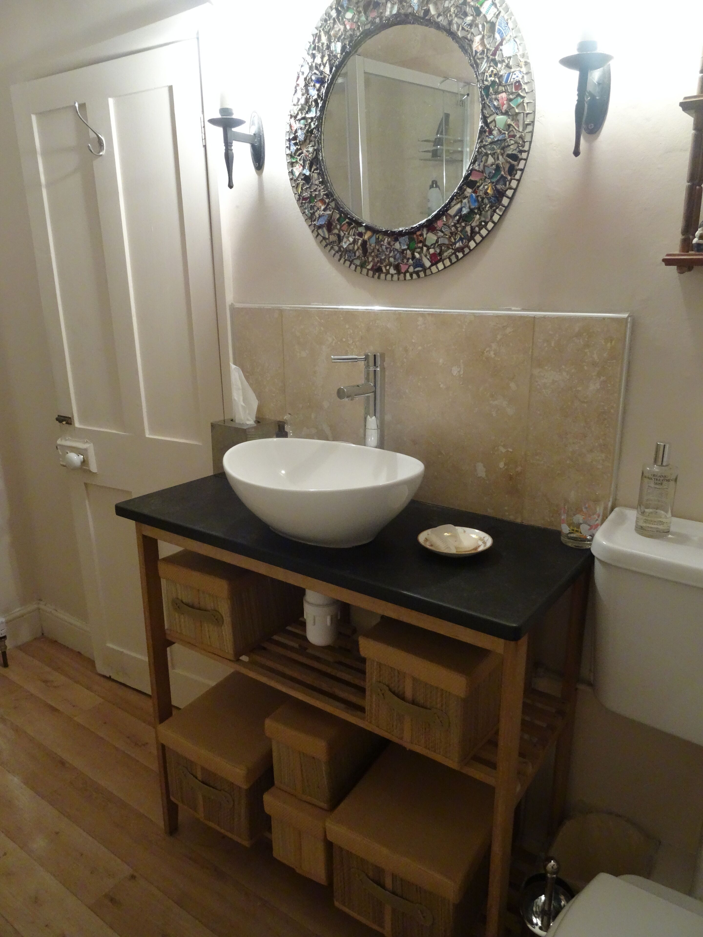 A bathroom with a white sink, a round mirror with a mosaic frame, and a wooden vanity with drawers.