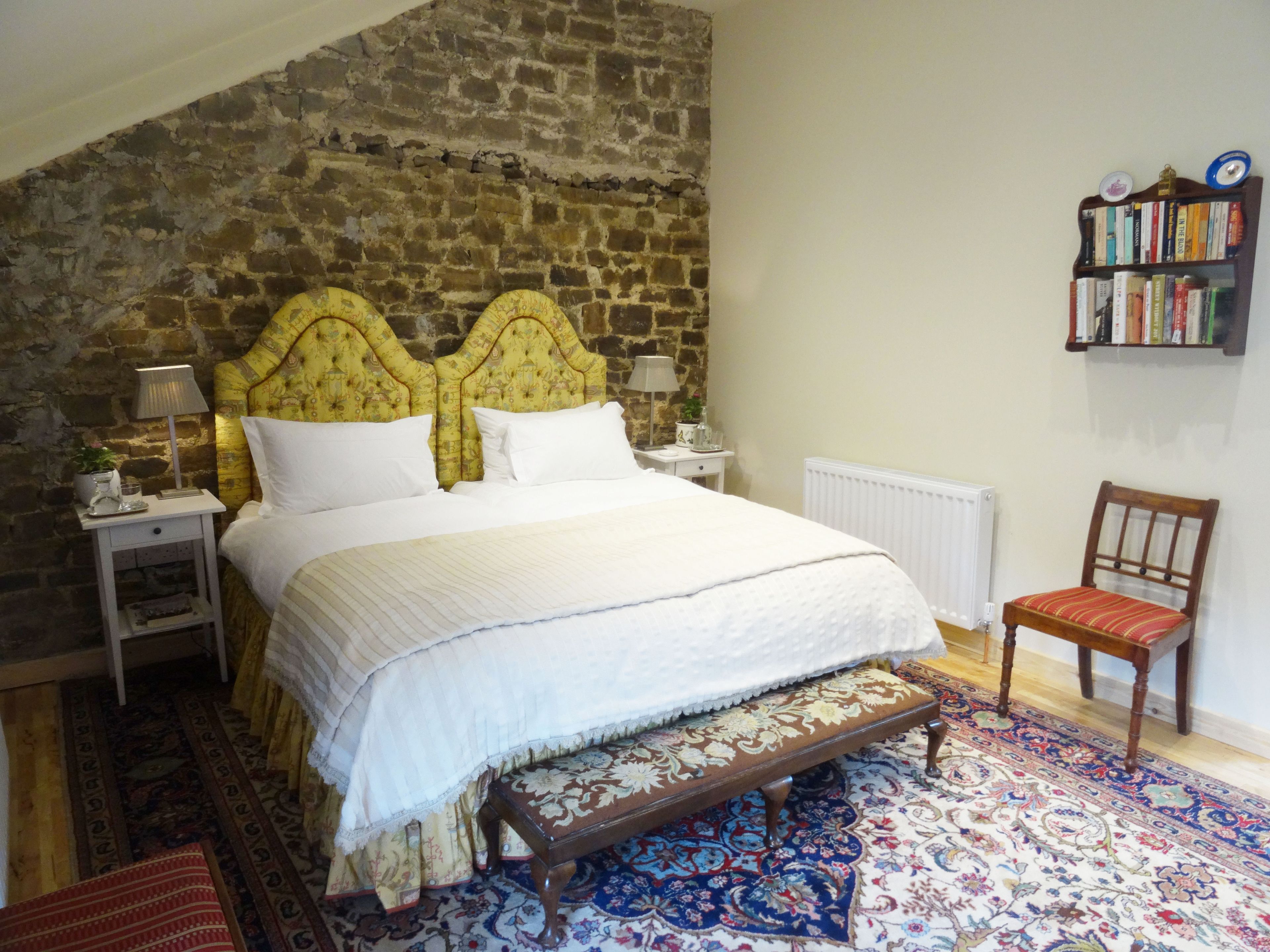 A bedroom with a stone wall, a wooden headboard, a white bed, a wooden bench, a patterned rug, and a bookshelf.