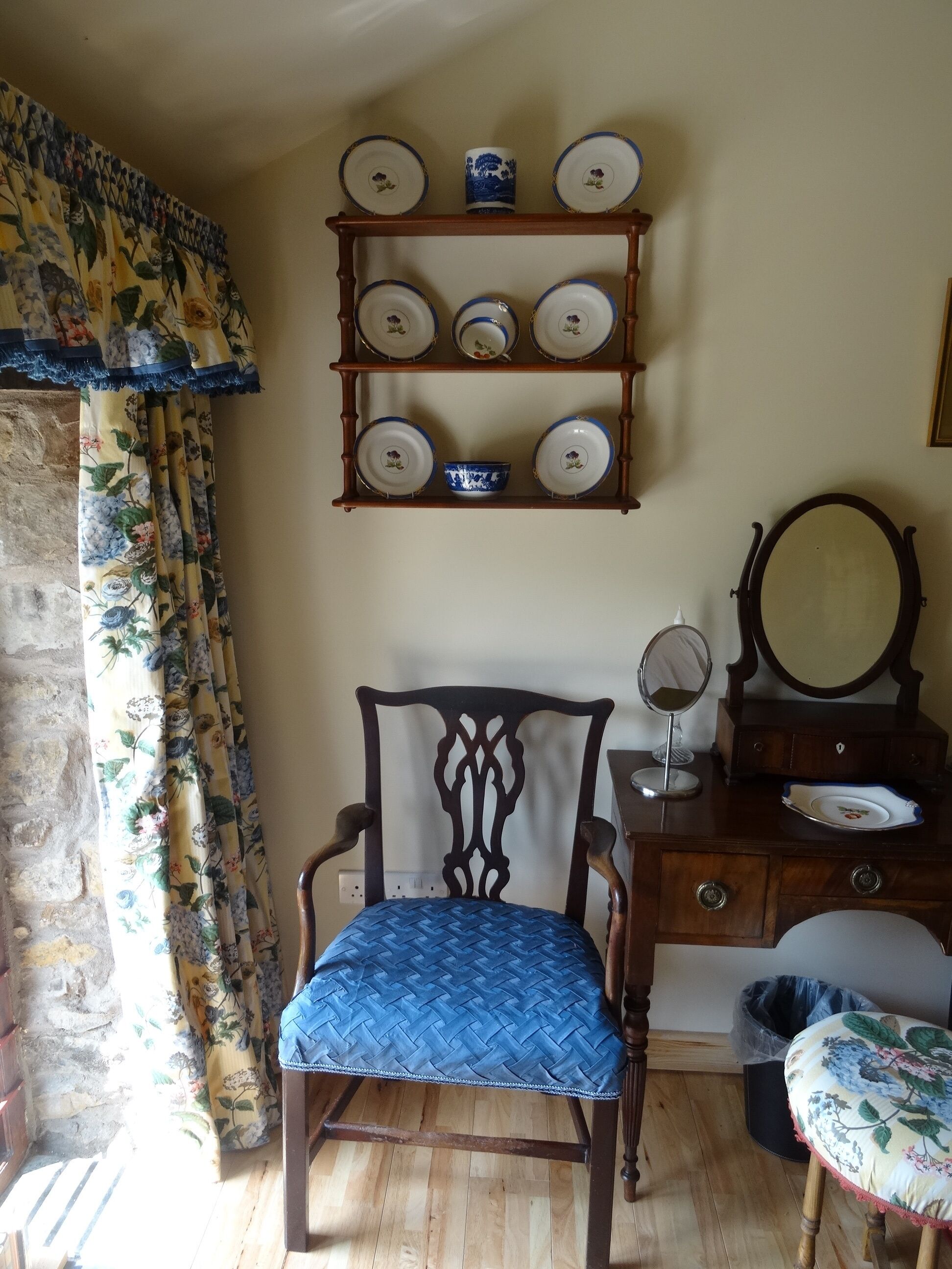 A room with a wooden chair with a blue cushion, a wooden dresser with a mirror, and a wall-mounted shelf with plates and a bowl.