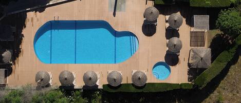 An aerial view of a swimming pool with a wooden deck, surrounded by lounge chairs and umbrellas.