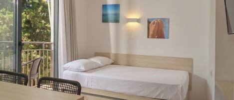 A single bed with a wooden headboard, a small table, and chairs, with a view of trees through a large window.