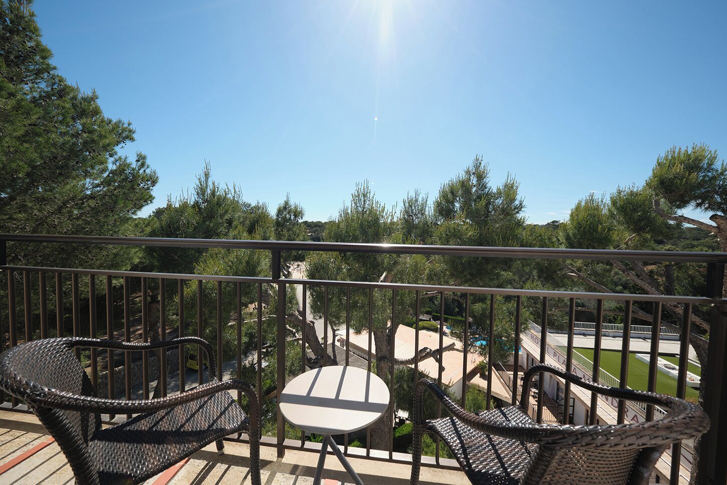 Un balcón con mobiliario de mimbre, una pequeña mesa y vistas a una piscina y árboles.