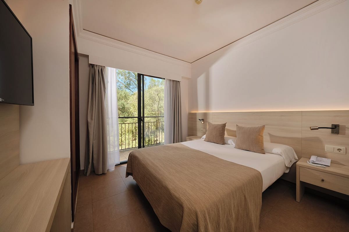 A hotel room with a large bed, a bedside table with a lamp, a flat-screen TV mounted on the wall, and a balcony with curtains.