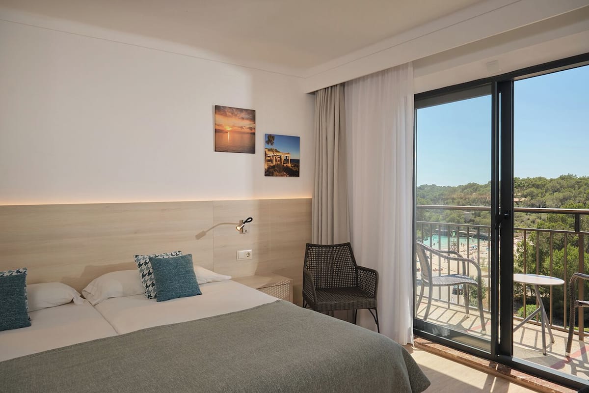 A hotel room with a bed, a chair, a balcony with a view of the beach and trees.