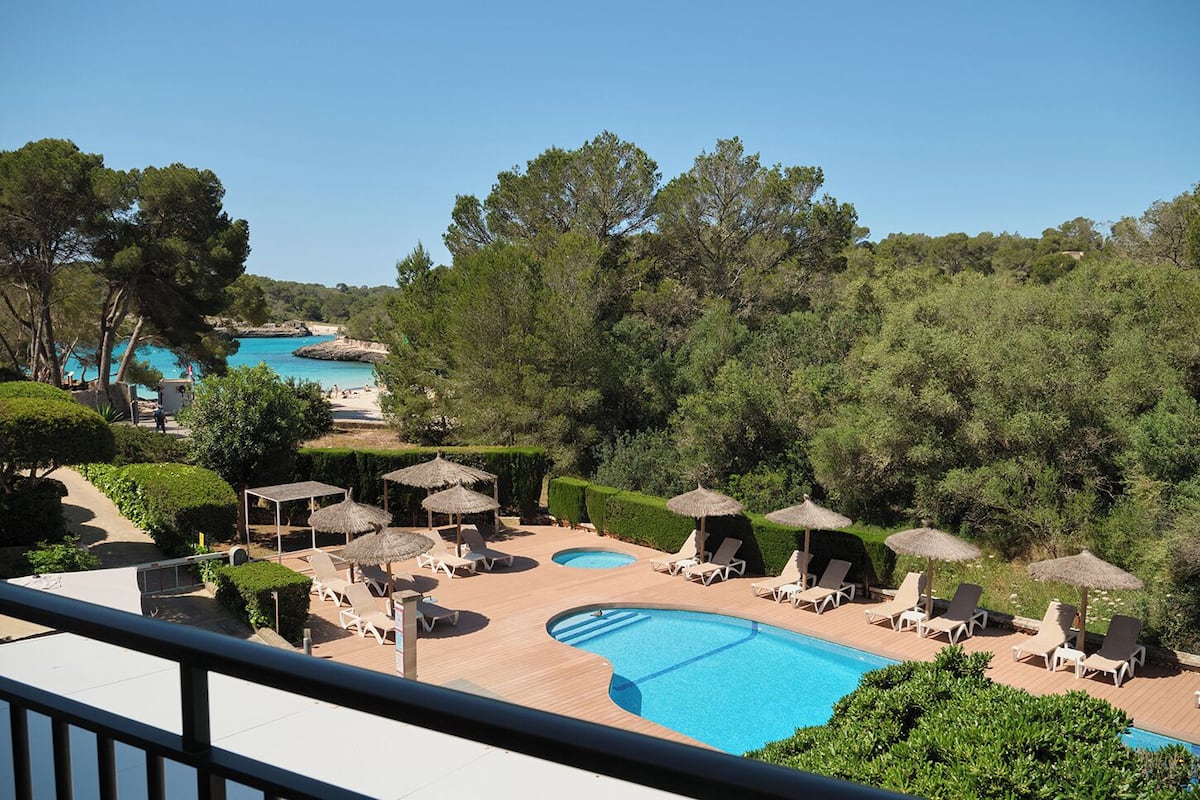 A pool area with lounge chairs and umbrellas, surrounded by greenery and overlooking a beach.