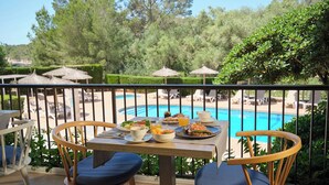 Uma mesa de jantar com pratos de comida, uma chávena e um copo de sumo de laranja numa varanda com vista para uma piscina e espreguiçadeiras.