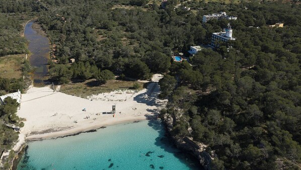 An aerial view of a coastal area with a beach, clear turquoise waters, and dense forested hills. - Hotel Playa Mondrago (Santanyi)