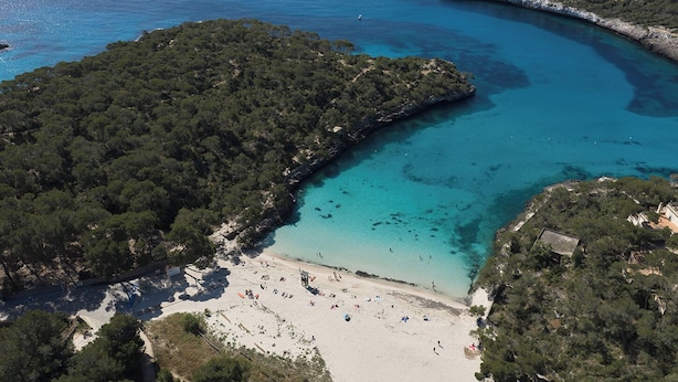 Vista aérea de uma área costeira com praia, águas turquesa cristalinas e colinas verdejantes e densas.