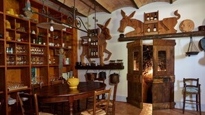 A rustic dining area with a wooden table and chairs, surrounded by shelves filled with glassware and bottles. A fireplace with a log fire is visible in the background.