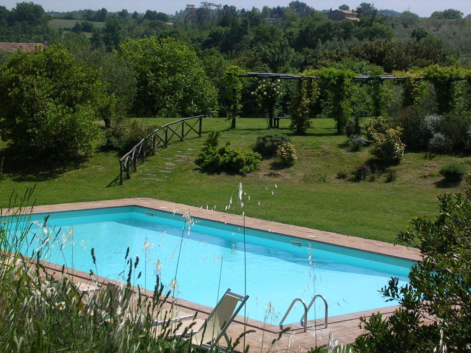 Piscina com deck de madeira e espreguiçadeiras, cercada por vegetação exuberante e uma parede de pedra.