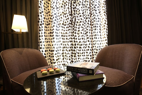 A room with two brown chairs, a table with a book titled "Book of Houses" and macarons, and a patterned curtain. - Lancaster Plaza Beirut (Beirut)