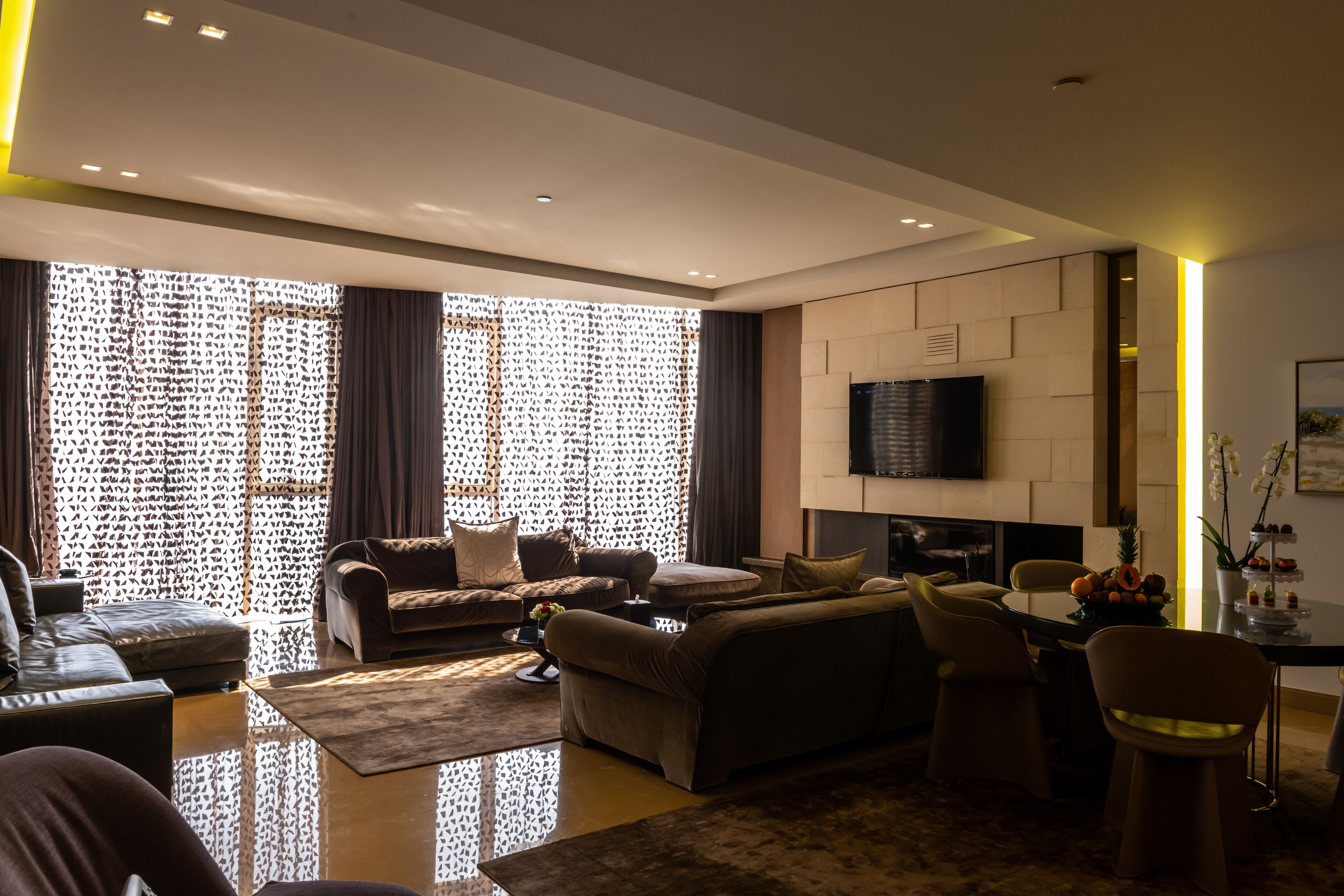 A modern living room with a sofa set, a flat-screen TV mounted on the wall, and a dining area with a table and chairs.