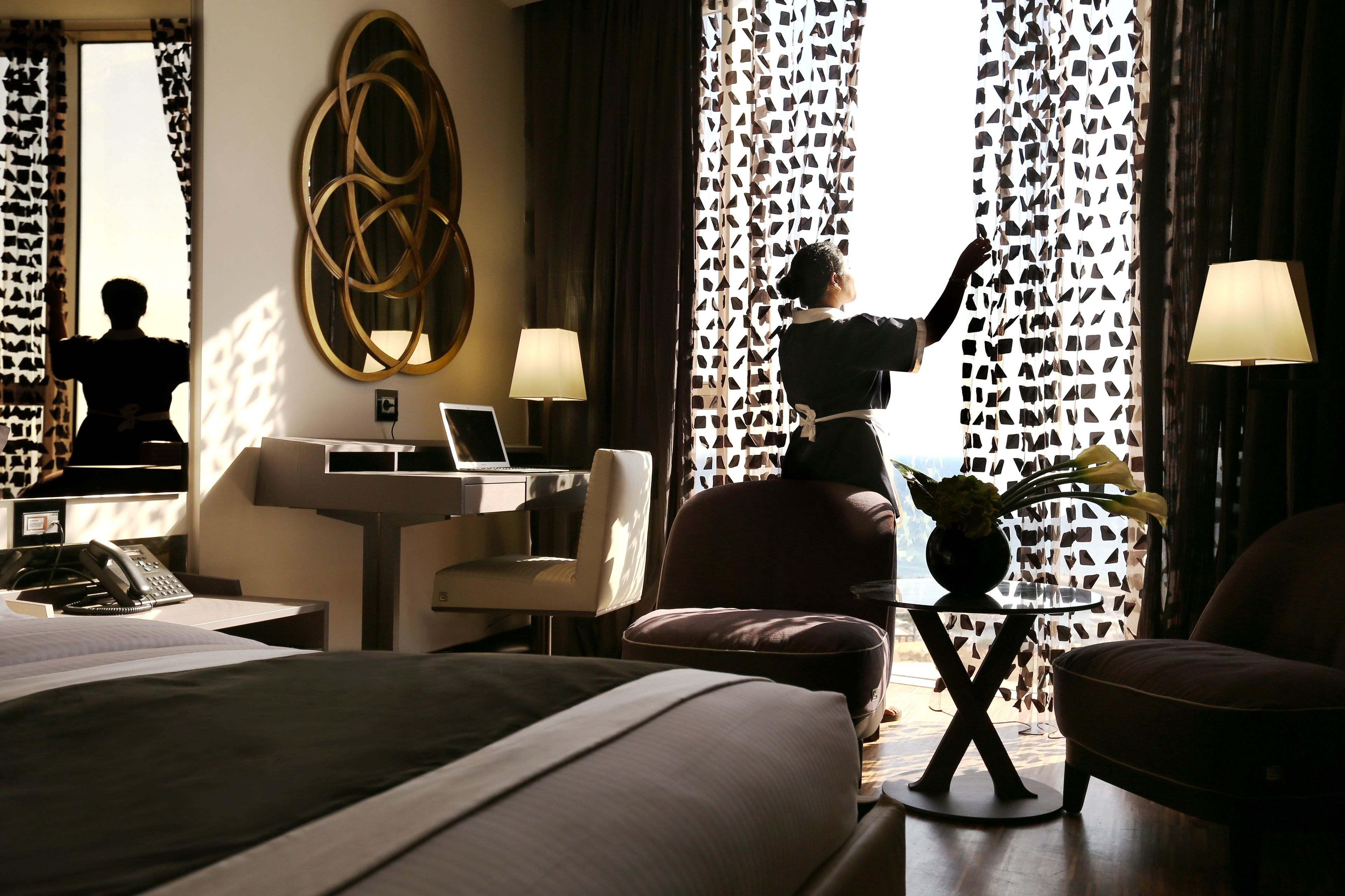 Una habitación de hotel con una cama, un escritorio con computadora, una silla, una lámpara y vistas al exterior a través de cortinas estampadas.