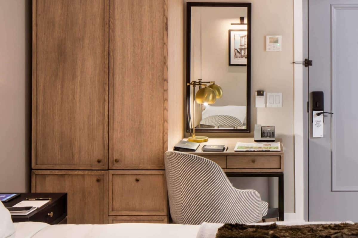 A hotel room with a wooden wardrobe, a desk with a lamp, a chair, a bed, and a door with a lock.