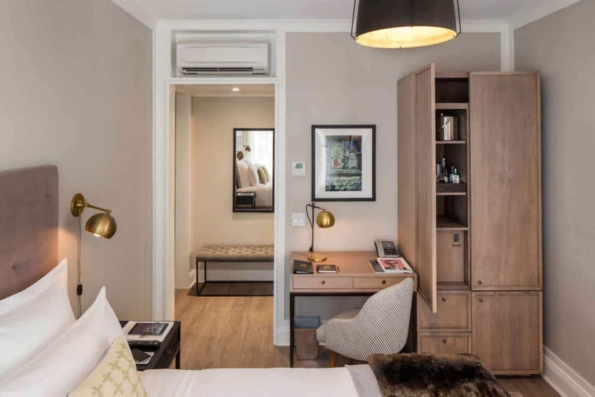 A hotel room with a bed, desk, chair, wardrobe, and an air conditioning unit.
