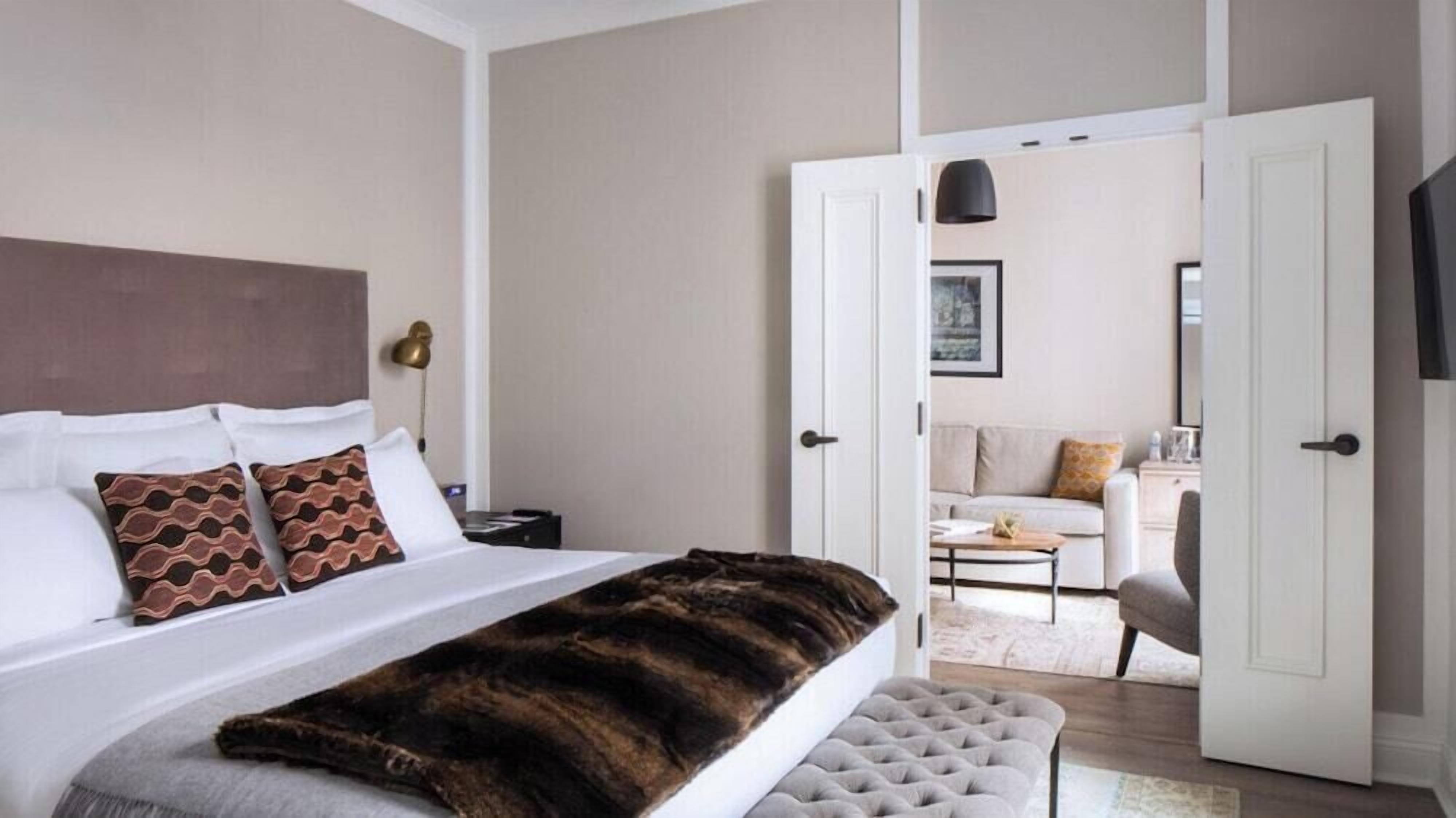 Quarto de hotel com cama, um banco, mesa de cabeceira, abajur e porta aberta para uma sala de estar.
