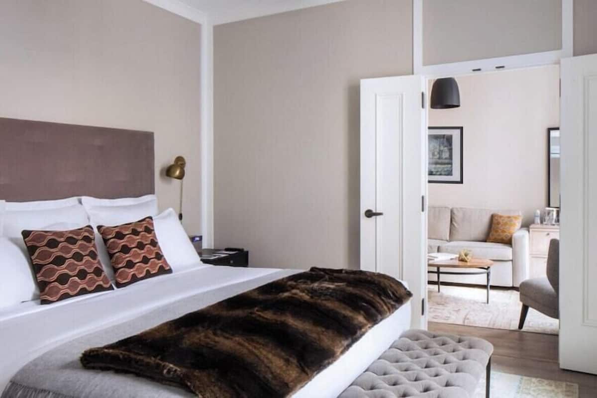 A hotel room with a bed, a bench, a nightstand, a lamp, and an open door leading to a living area.