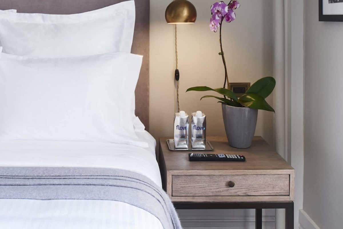 A neatly made bed with white linens, a bedside table with a remote control, a potted orchid, and two bottles labeled 'flow' on a wooden nightstand.