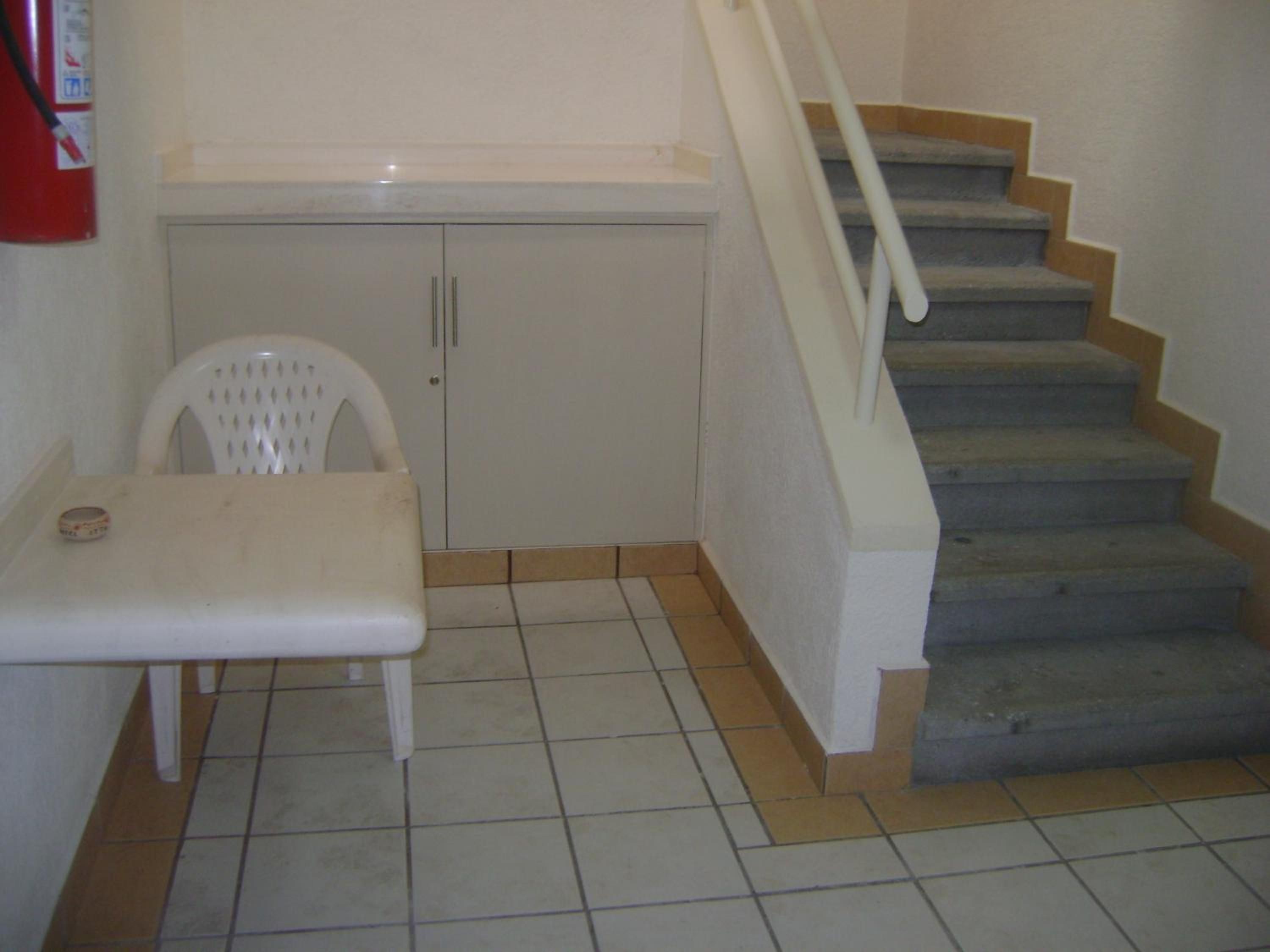 A corner of a room with a white chair, a small table, a cabinet, and a staircase with a handrail.