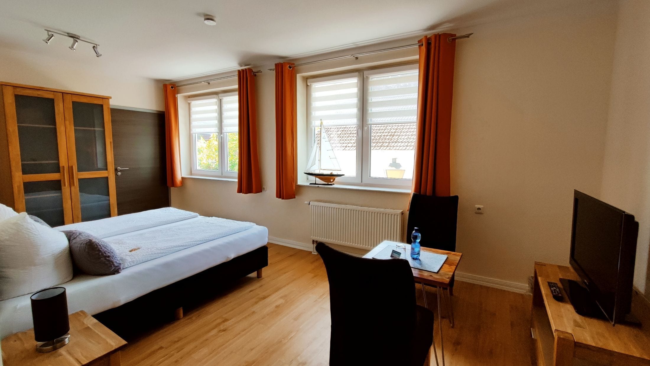 Quarto de hotel com cama, escrivaninha, cadeira, televisão e janela com cortinas.