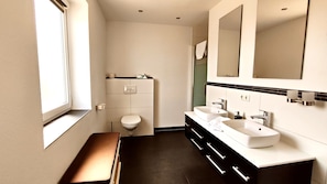 Een moderne badkamer met toilet, twee wastafels, een spiegel en een raam.