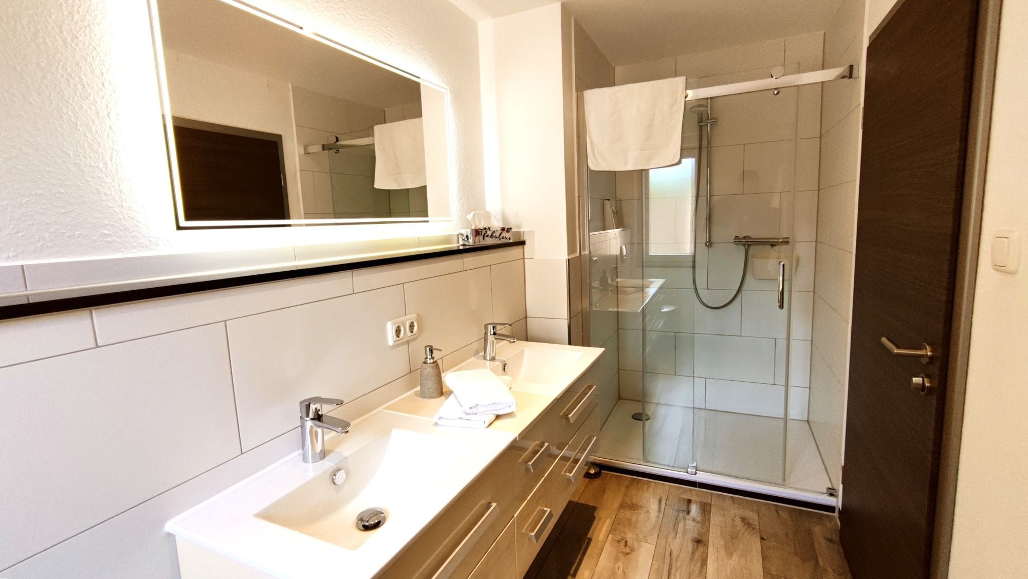 A modern bathroom with a large mirror, a sink with a vanity, a glass shower enclosure, and a wooden floor.