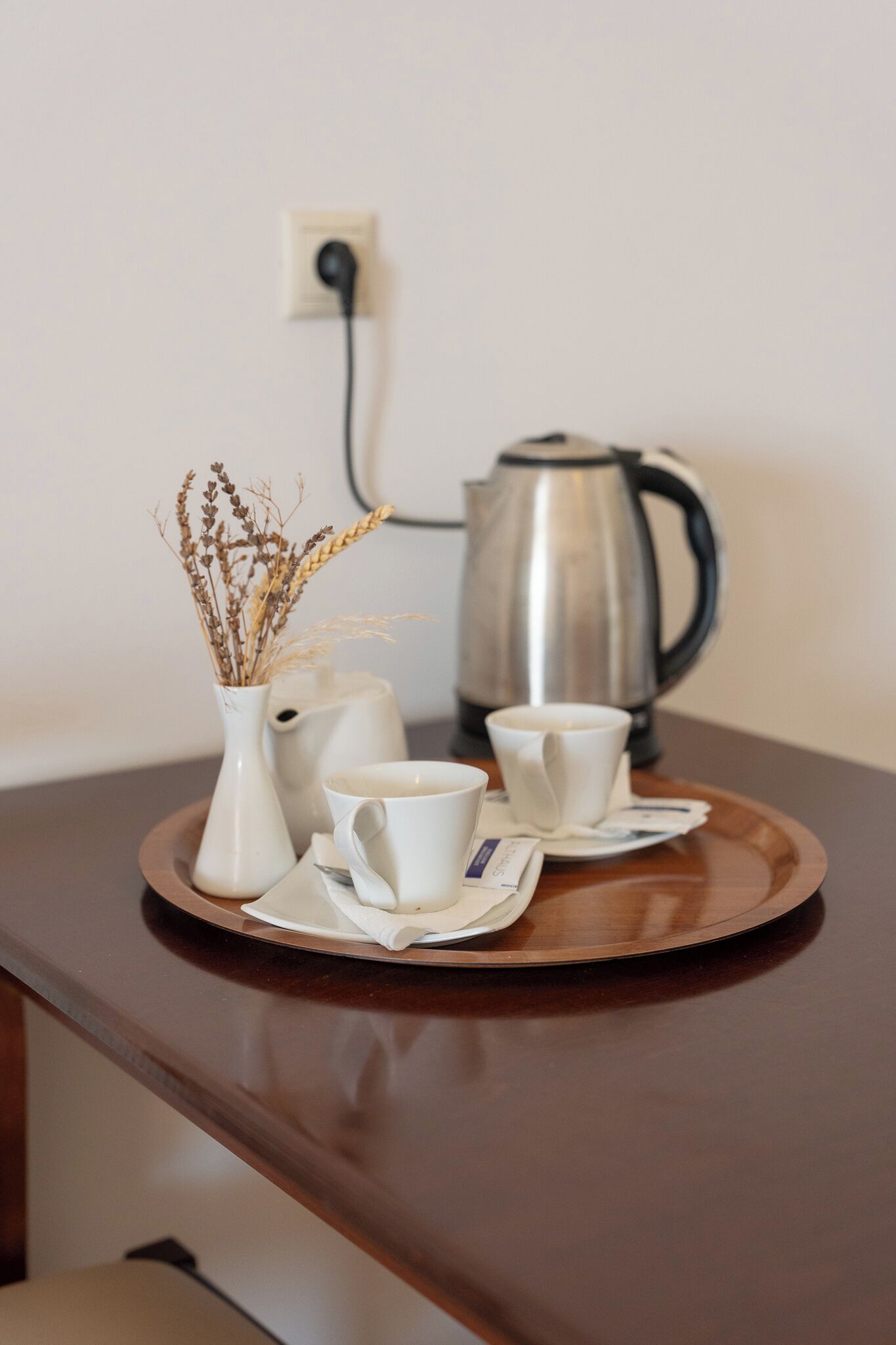 A tray with a white vase, two white cups, and a silver coffee pot on a wooden table.