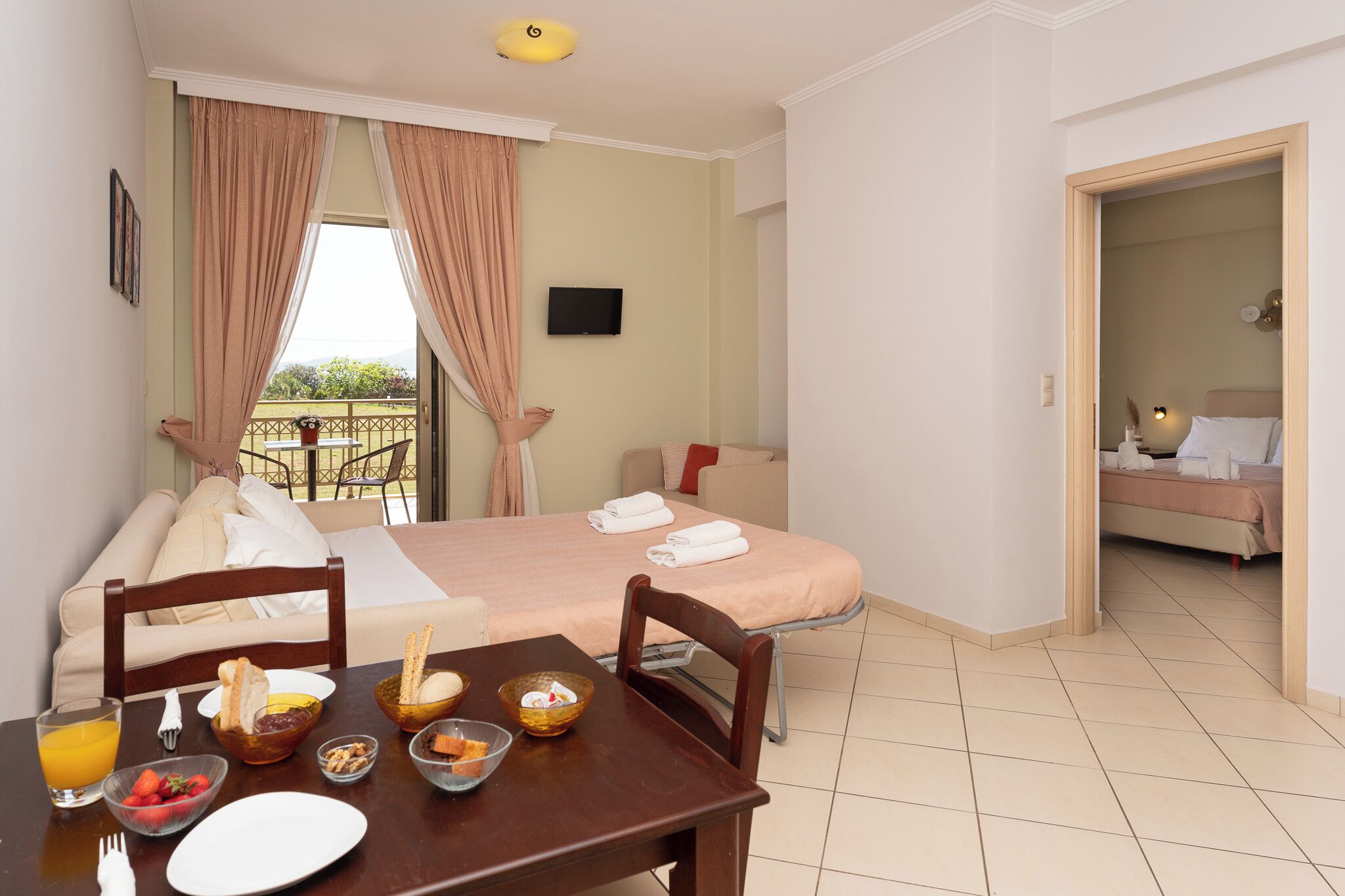 A hotel room with a dining area, a bed, a TV, and a balcony with curtains.