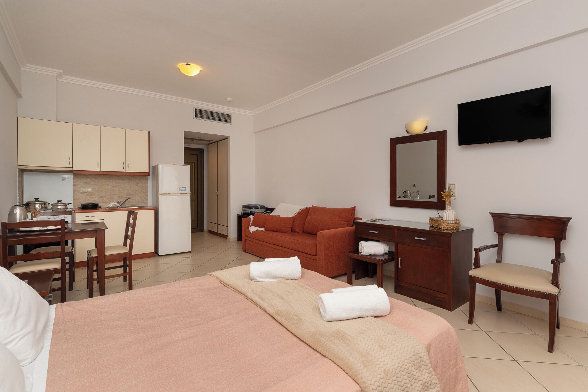 Une chambre d’hôtel comprenant un lit, un bureau, une chaise, une télévision, un miroir, un coin cuisine et une table à manger.