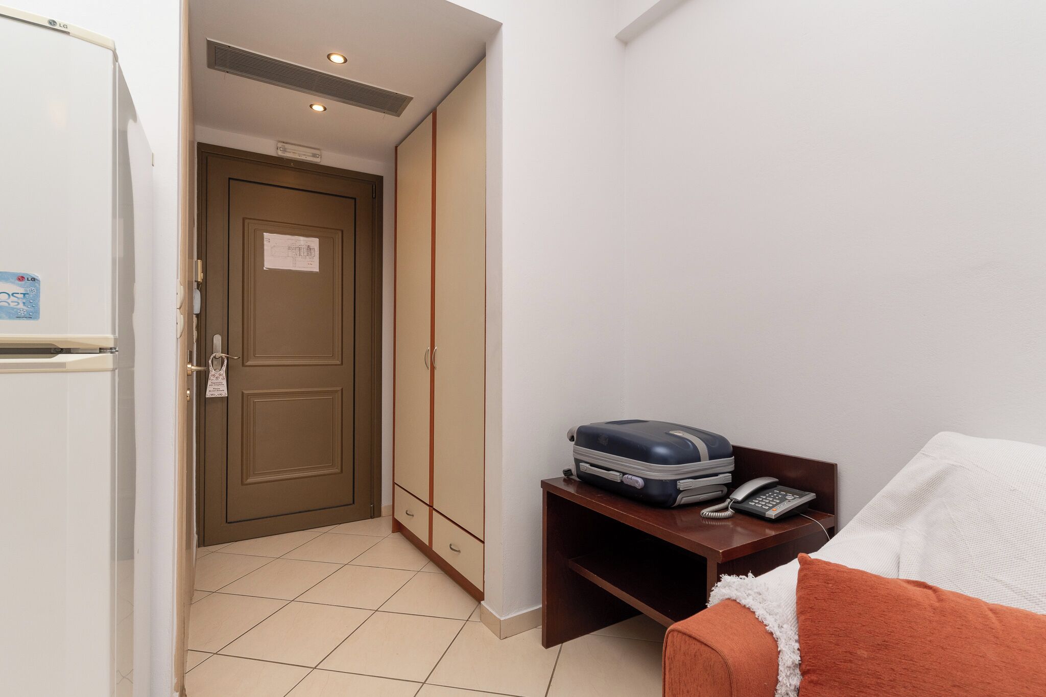 A hotel room with a bed, a bedside table with a phone and a suitcase, a wardrobe, and a door with a sign.