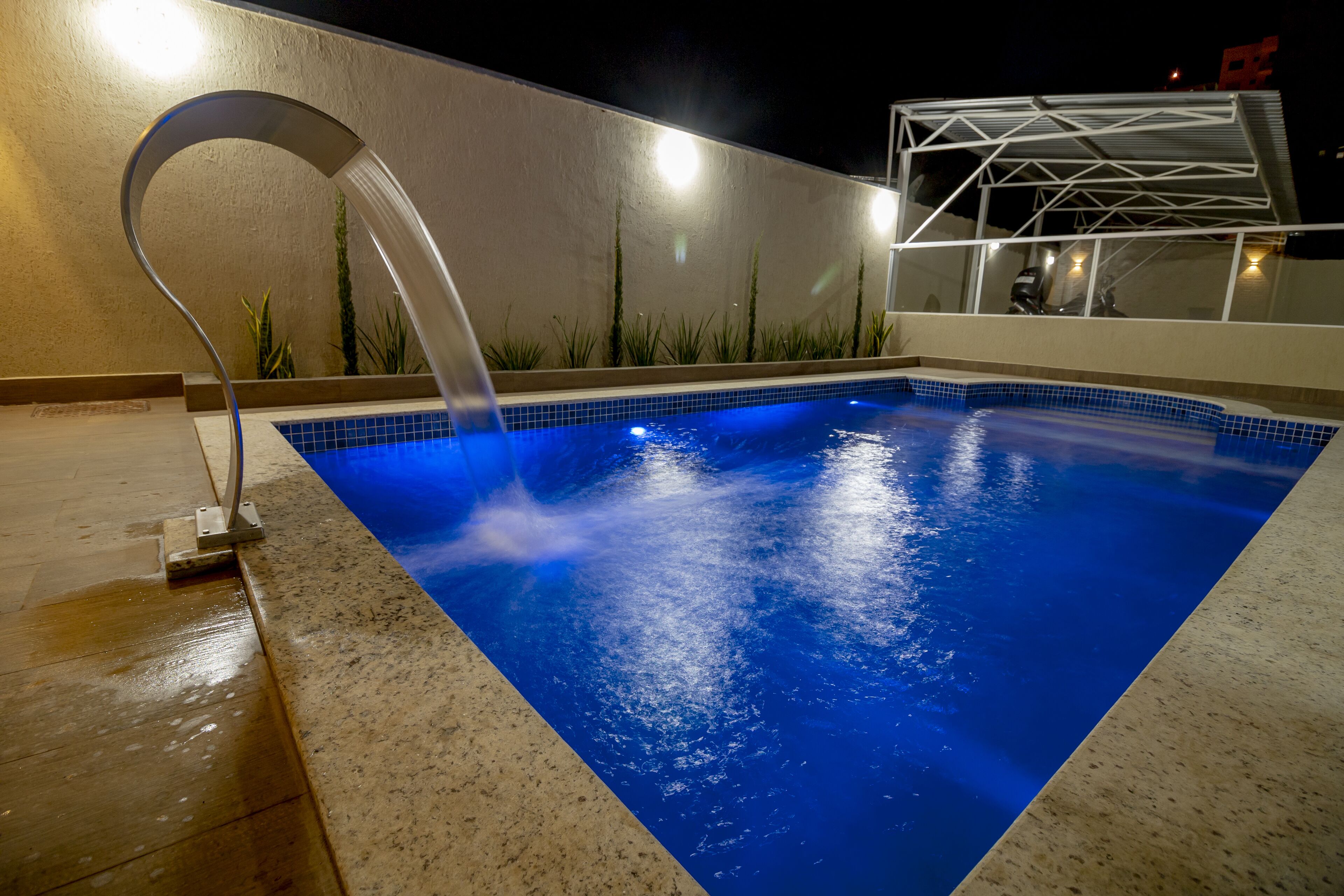 A swimming pool with a water feature, surrounded by a tiled deck and a low wall with plants.
