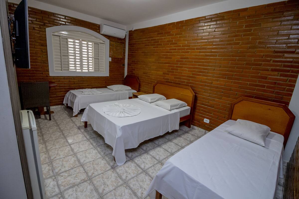 A room with three single beds, a wooden brick wall, a window with shutters, and a flat-screen TV mounted on the wall.