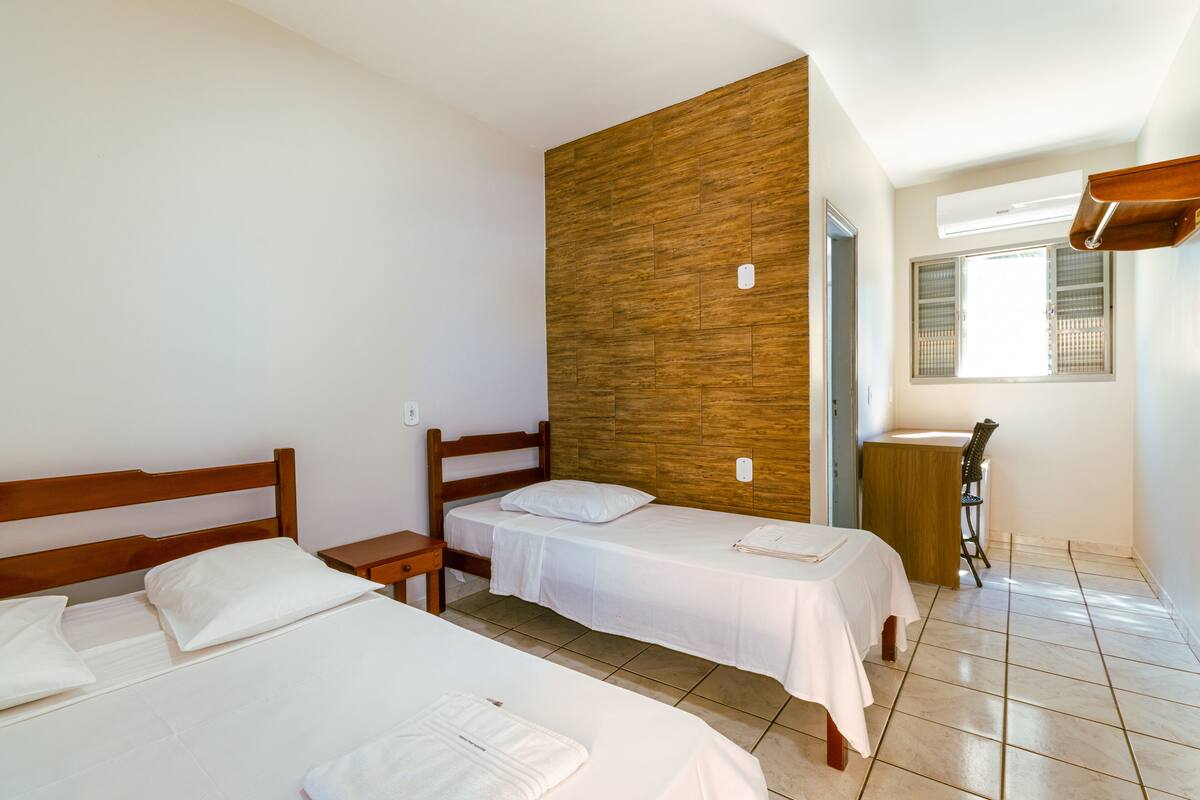 A hotel room with two beds, a wooden accent wall, a desk with a chair, and a window with blinds.