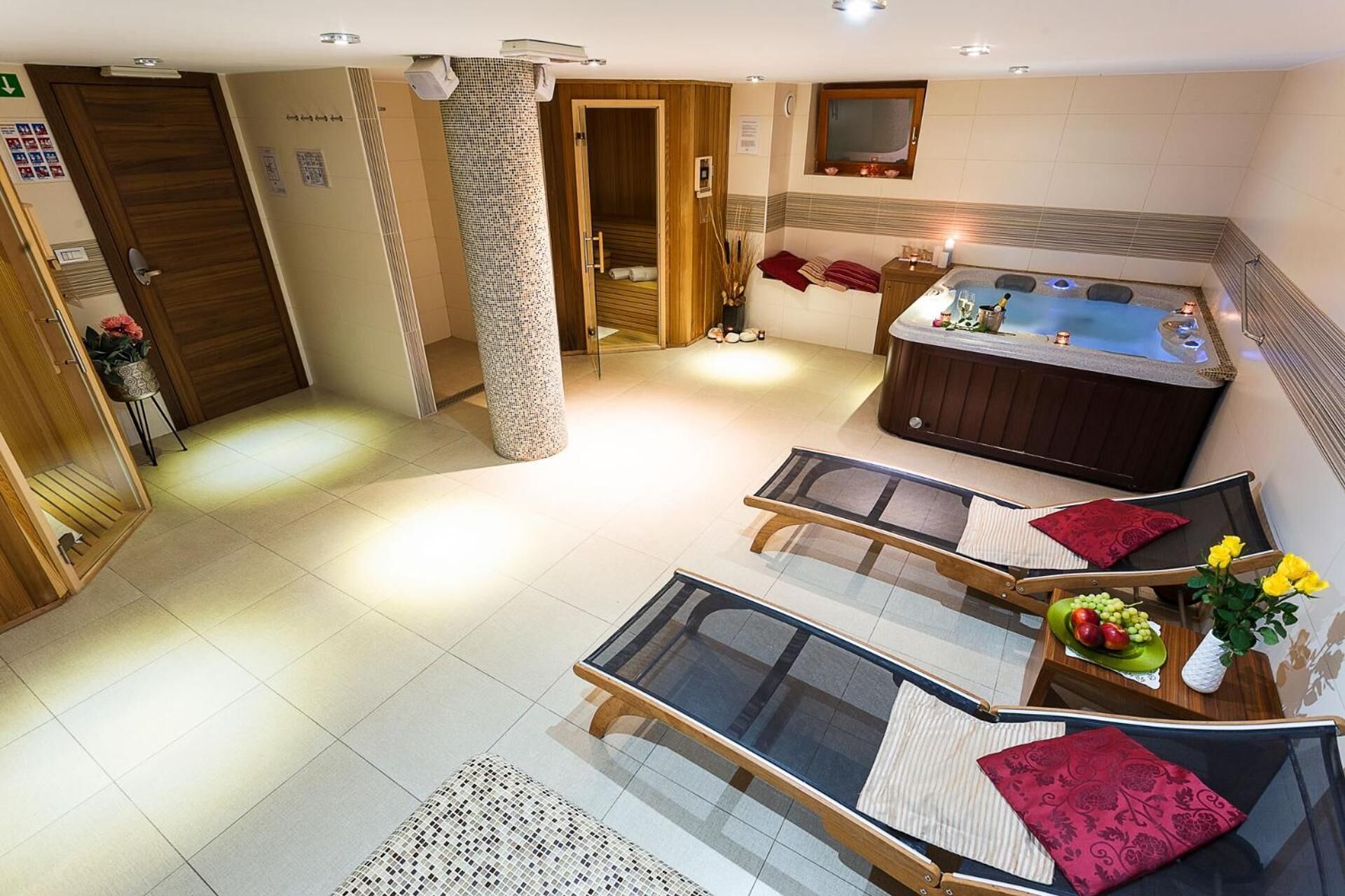 A modern hotel room with a jacuzzi, two lounge chairs, a small table with a fruit bowl, and a decorative pillar.