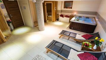 A modern hotel room with a jacuzzi, two lounge chairs, a small table with a fruit bowl, and a decorative pillar.