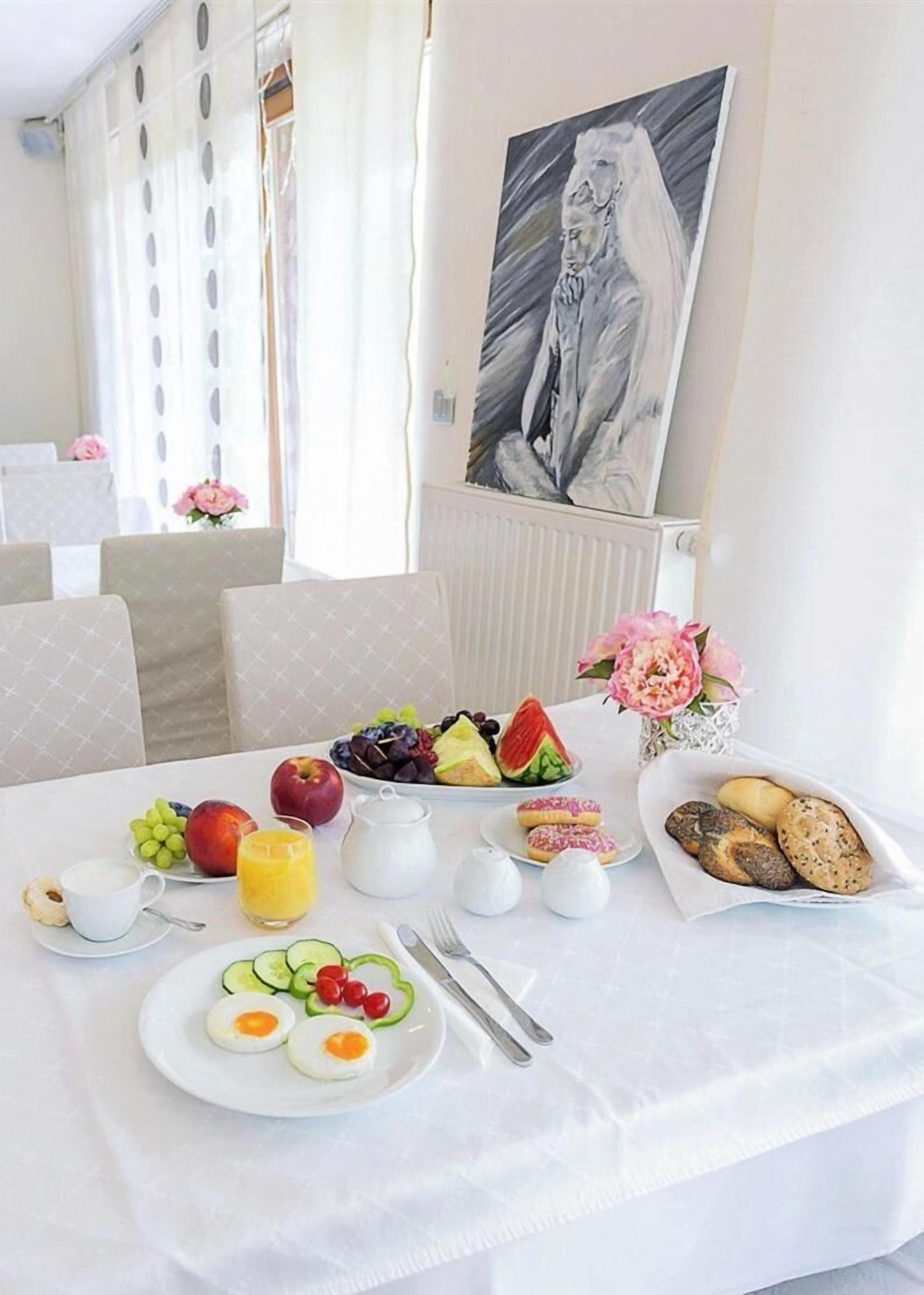 Una mesa de desayuno con frutas, pan y huevos, con un cuadro y flores al fondo.