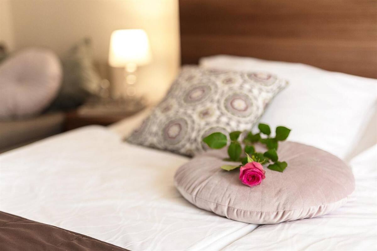 Un lit bien fait, agrémenté d’une seule rose posée sur un coussin rond, d’un autre coussin à motifs et d’une lampe en arrière-plan.