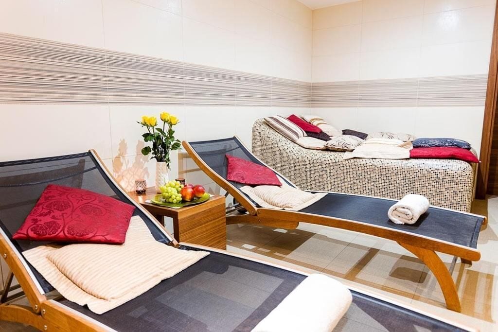 Un salón de spa con dos camillas de masaje, una pequeña mesa de madera con un jarrón de flores, y un sofá estampado.