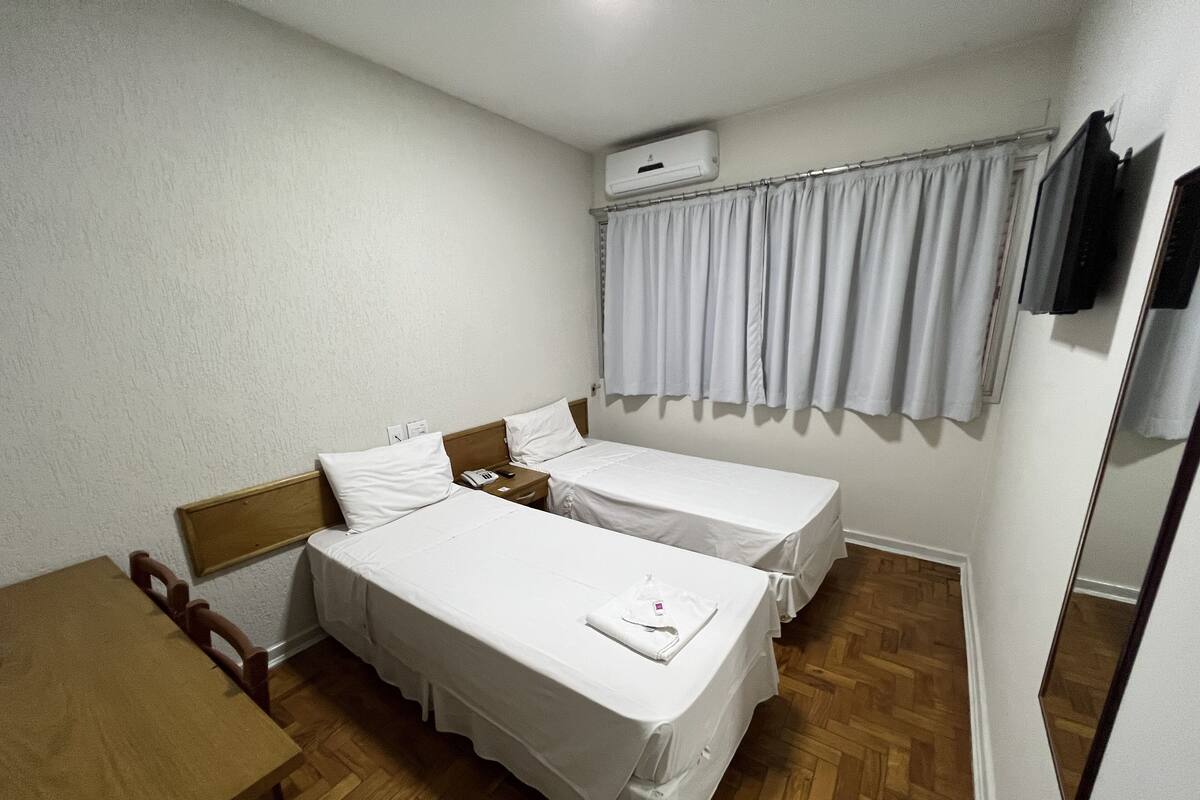 A hotel room with two single beds, a wooden desk, a chair, a mirror, and a window with curtains.