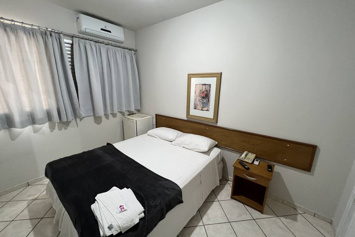 A hotel room with a bed, a bedside table with a phone, a small cabinet, a window with curtains, and an air conditioning unit on the wall.