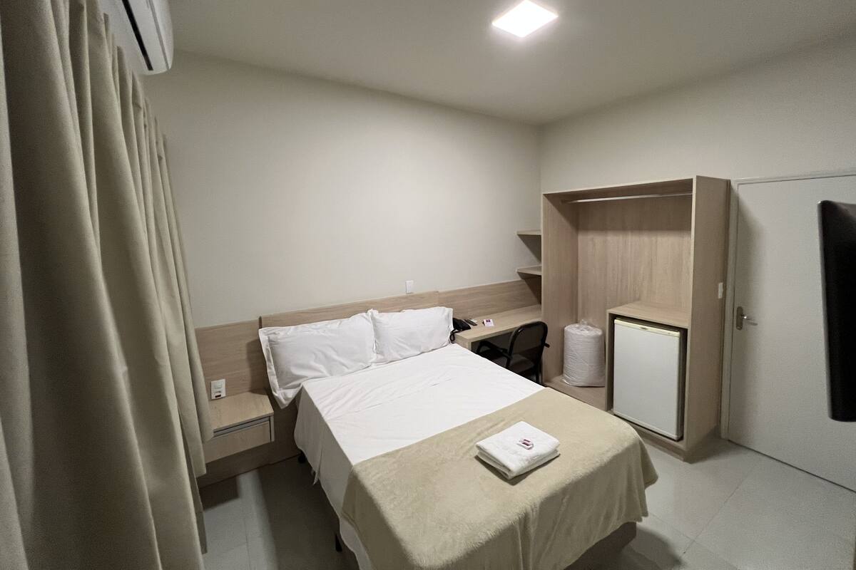 A hotel room with a bed, bedside table, a desk with a chair, a wardrobe, and a toilet.