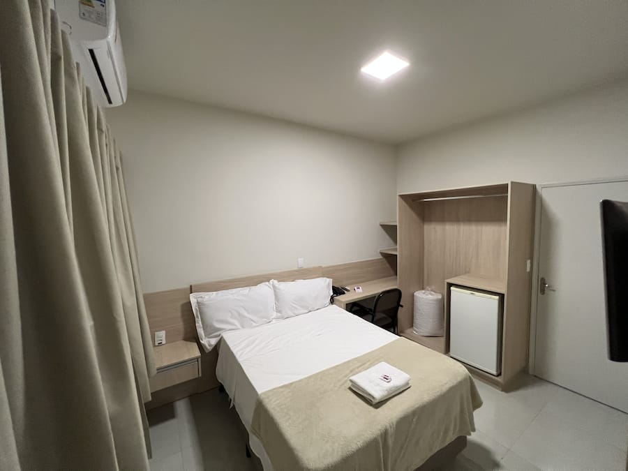 A hotel room with a bed, bedside table, a desk with a chair, a wardrobe, and a toilet.