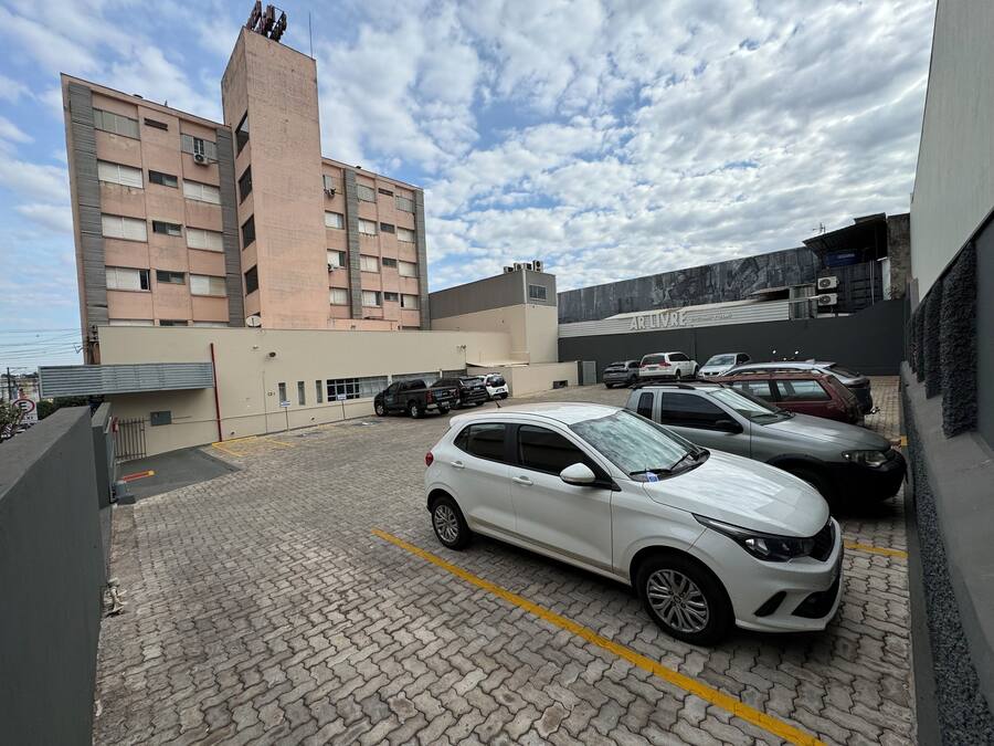 A parking area with several cars, a building with a sign reading "AR LIVRE," and a large brick building in the background.