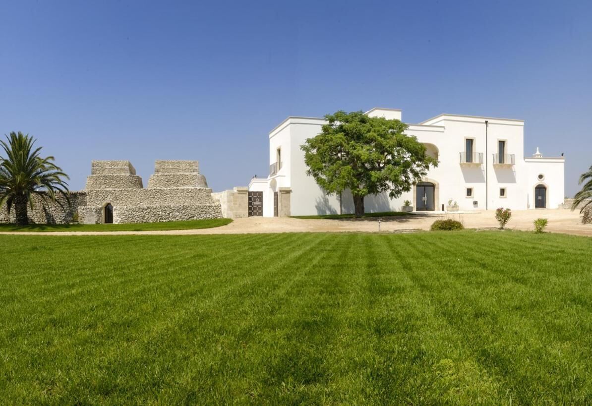 Un edificio blanco con un gran árbol en primer plano, una estructura de piedra a la izquierda y un césped cuidado.