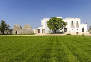 Un edificio blanco con un gran árbol en primer plano, una estructura de piedra a la izquierda y un césped cuidado.
