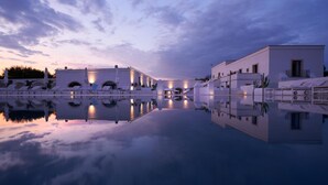 Un hotel moderno con piscina que refleja el cielo, edificios blancos y sillones de descanso.