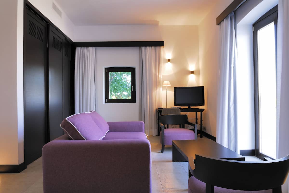A modern hotel room with a purple sofa, a small table, a TV, and a window with curtains.