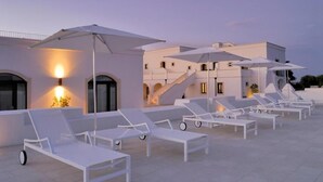 A rooftop lounge area with white sun loungers and umbrellas, a staircase, and a building with arched windows and warm lighting. - Masseria Bagnara (Lizzano)