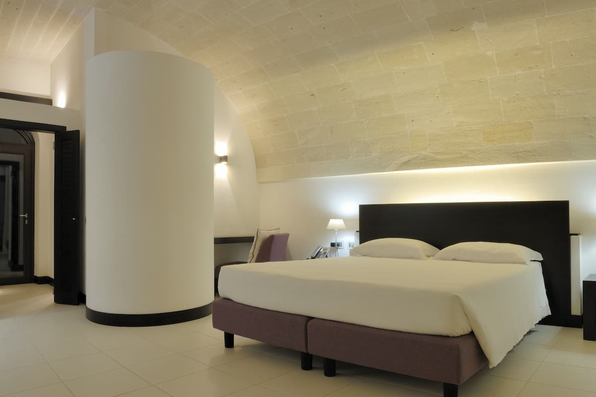 A modern hotel room with a large bed, two bedside tables with lamps, a curved wall, and a door leading to another room.
