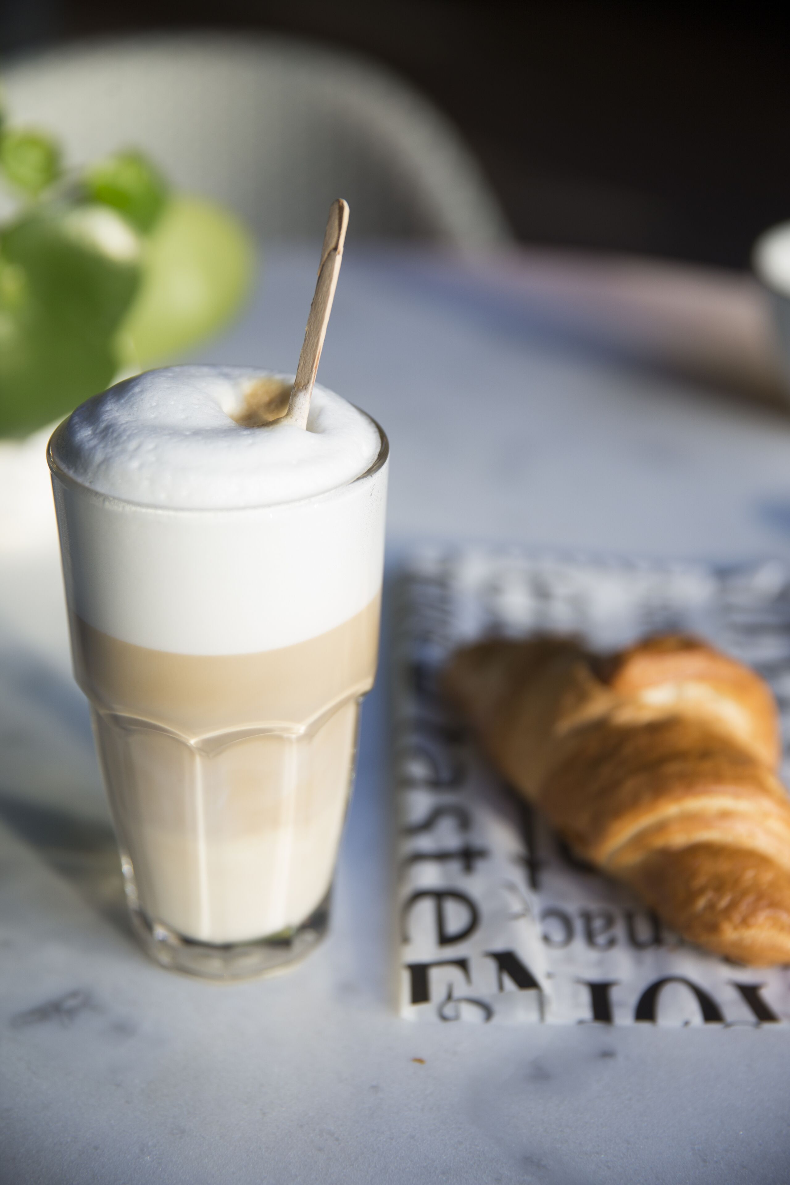 Um copo com bebida em camadas, um canudo, um croissant e um jornal sobre uma mesa.
